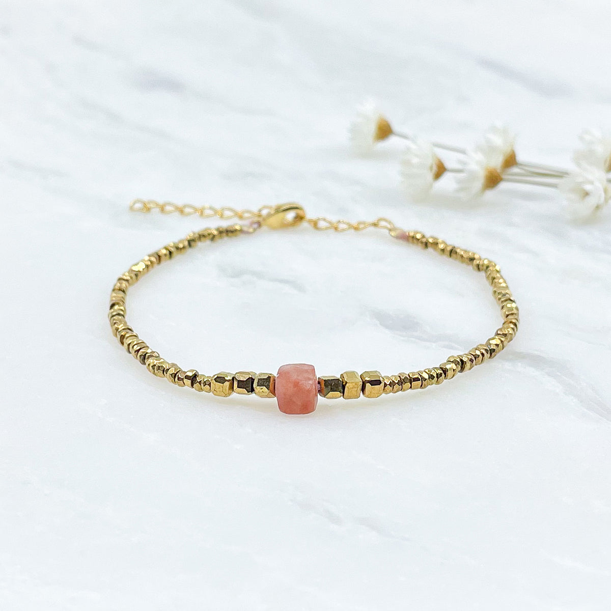 Bracelet mettant en lumière une perle cubique facettée de pierre fine naturelle de Jaspe rose pêche. Elle est entourée de perles d'hématites dorées.