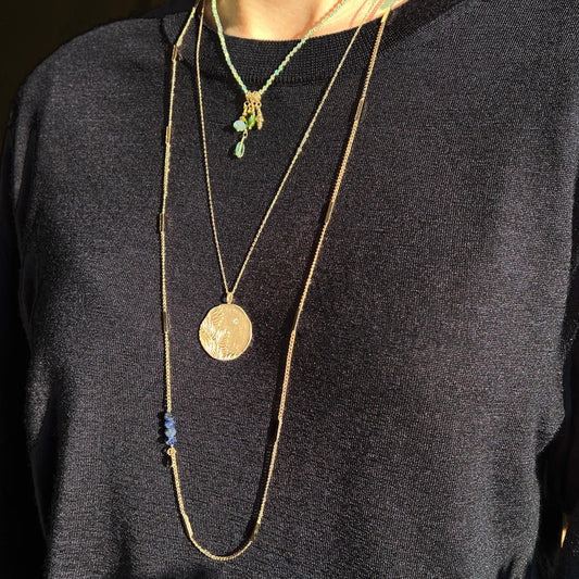 Long collier en chaîne maille forçat de 50 cm. Médaille de 2.6 cm de diamètre au contours irréguliers représentant de beaux feuillages et petit éclat scintillant en forme d'étoile. Or mat en partie recyclé couleur champagne