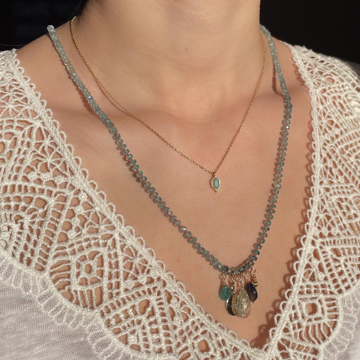 Collier long de 60 cm en pierres fines de zircon naturel d'un bleu azur. 5 charms : 3 pierres fines naturelles de teintes bleu-vert, une médaille ronde et un pendentif en petites perles plaqué or. Chaîne de réglage de 4 cm et fermoir plaqué or 18 carats