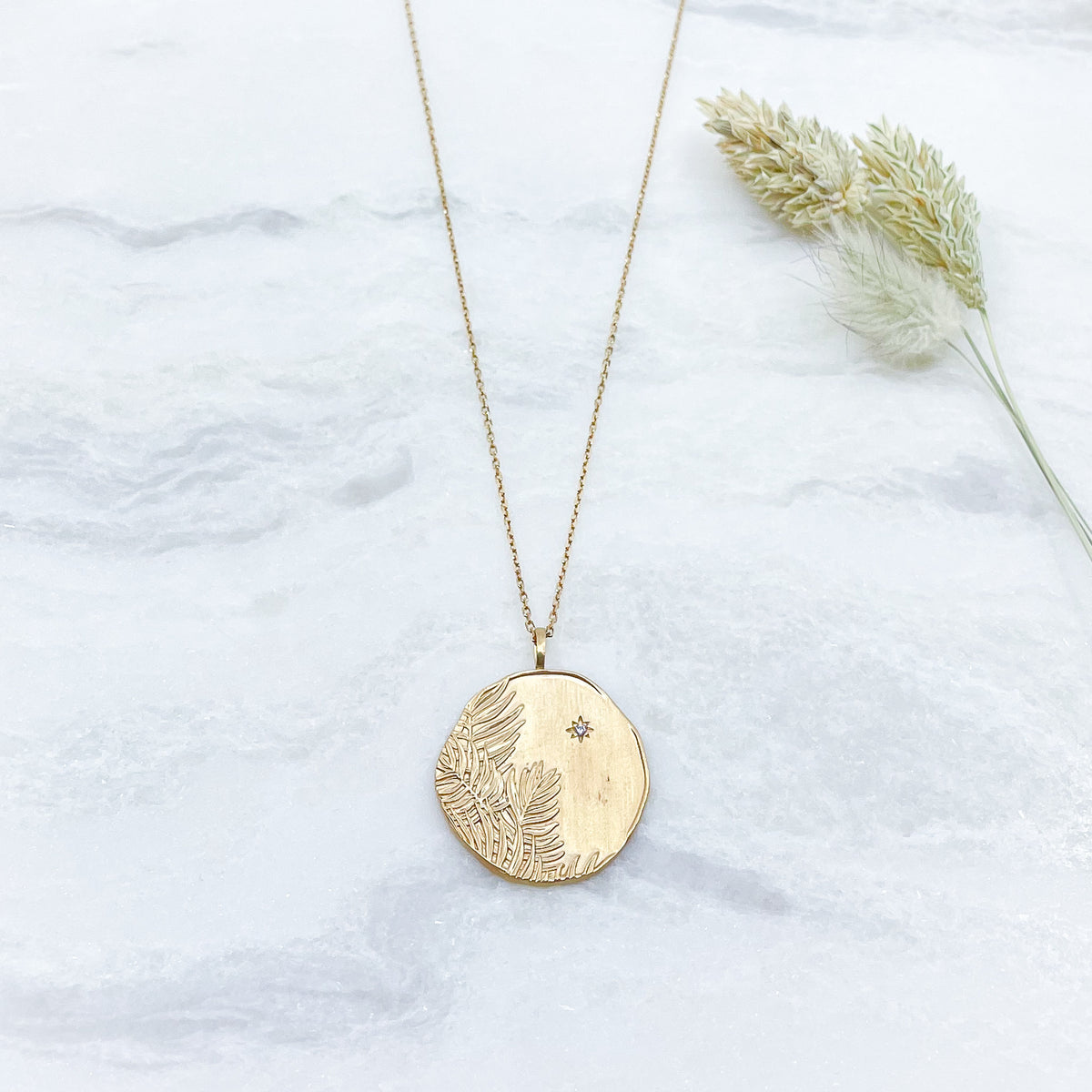 Long collier en chaîne maille forçat de 50 cm. Médaille de 2.6 cm de diamètre au contours irréguliers représentant de beaux feuillages et petit éclat scintillant en forme d'étoile. Or mat en partie recyclé couleur champagne