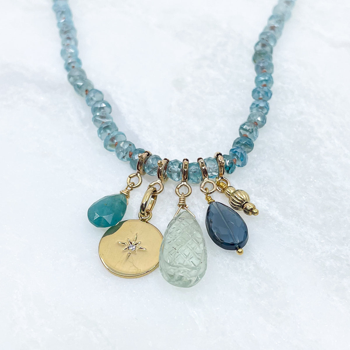 Collier long de 60 cm en pierres fines de zircon naturel d'un bleu azur. 5 charms : 3 pierres fines naturelles de teintes bleu-vert, une médaille ronde et un pendentif en petites perles plaqué or. Chaîne de réglage de 4 cm et fermoir plaqué or 18 carats