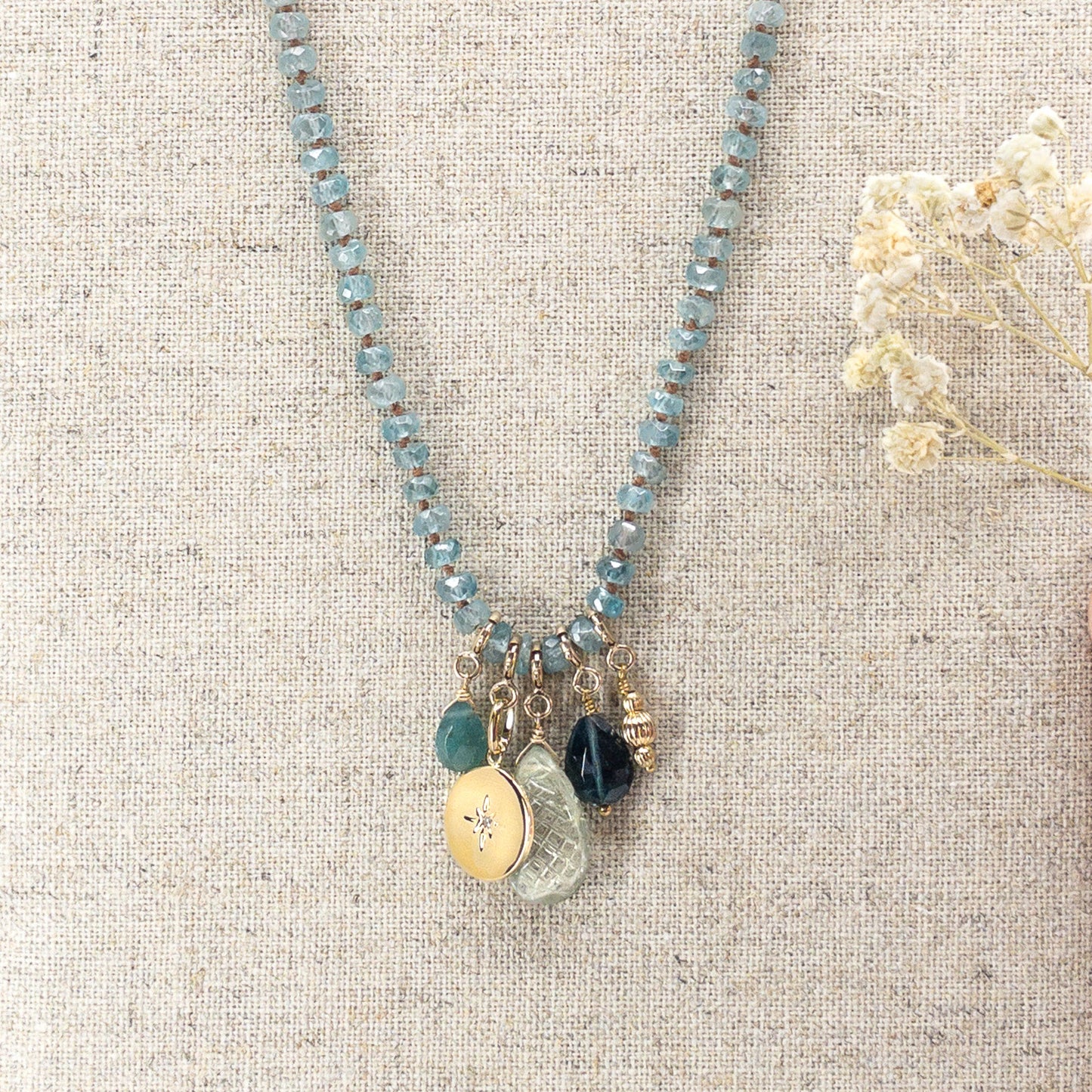 Collier long de 60 cm en pierres fines de zircon naturel d'un bleu azur. 5 charms : 3 pierres fines naturelles de teintes bleu-vert, une médaille ronde et un pendentif en petites perles plaqué or. Chaîne de réglage de 4 cm et fermoir plaqué or 18 carats