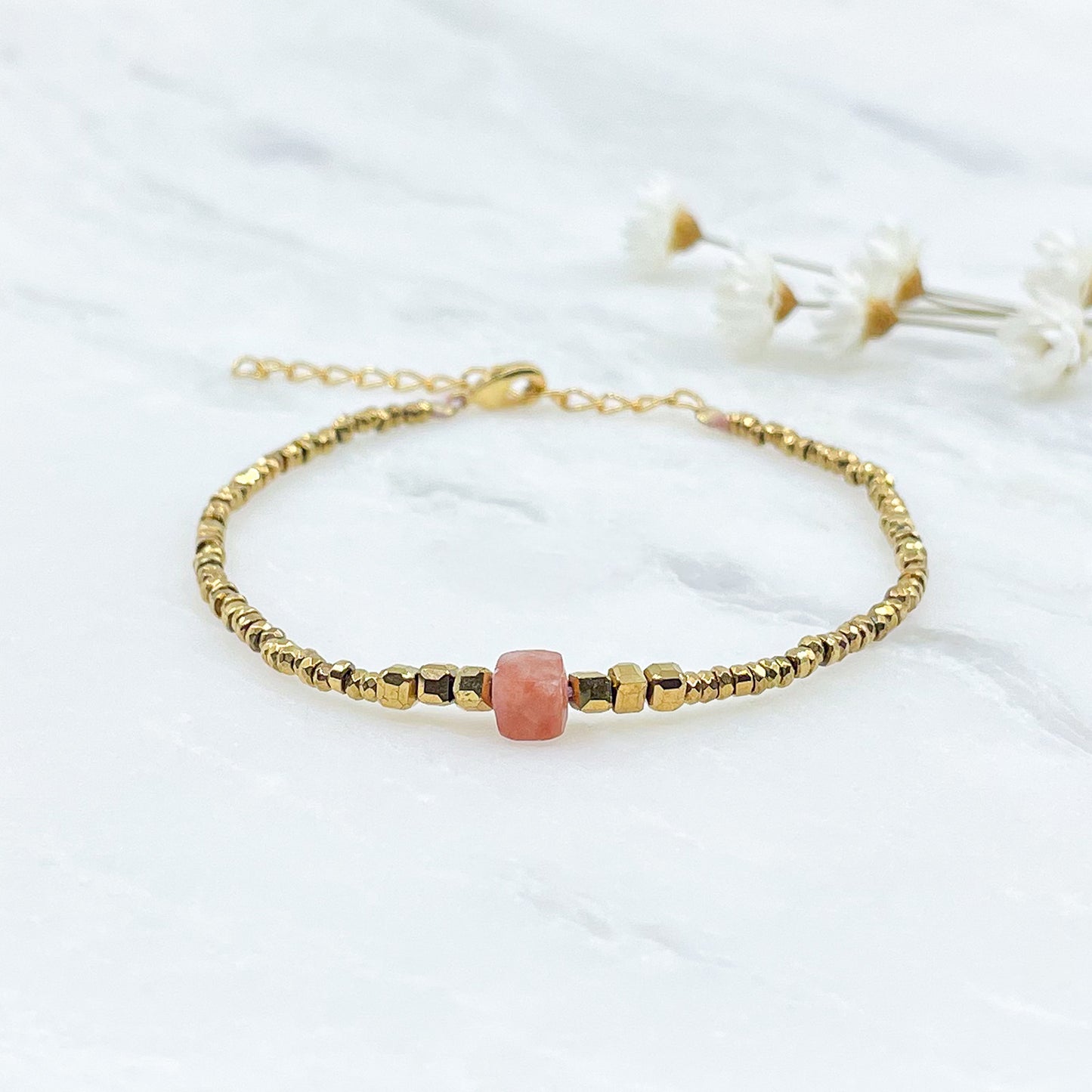 Bracelet mettant en lumière une perle cubique facettée de pierre fine naturelle de Jaspe rose pêche. Elle est entourée de perles d'hématites dorées. 