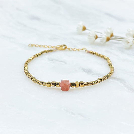 Bracelet mettant en lumière une perle cubique facettée de pierre fine naturelle de Jaspe rose pêche. Elle est entourée de perles d'hématites dorées. 