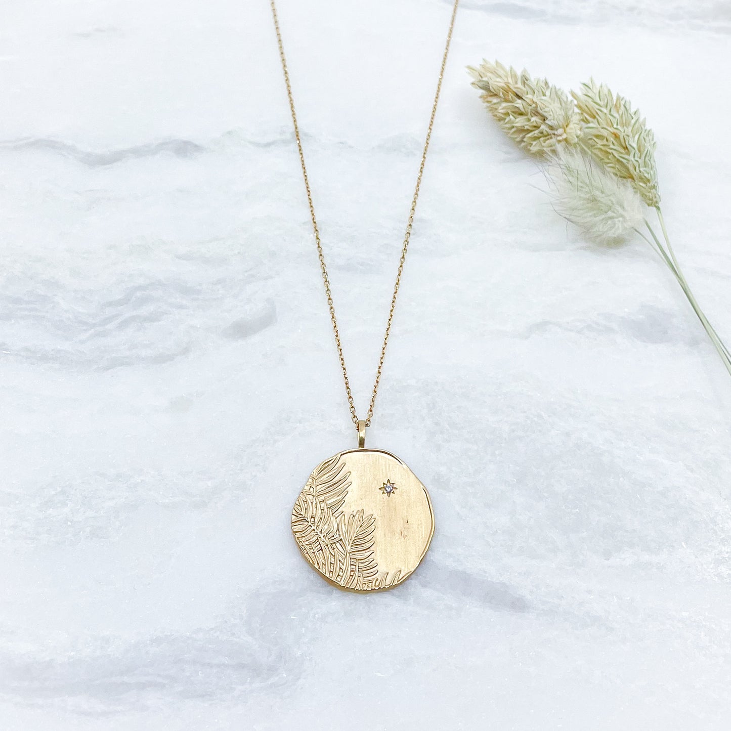 Long collier en chaîne maille forçat de 50 cm. Médaille de 2.6 cm de diamètre au contours irréguliers représentant de beaux feuillages et petit éclat scintillant en forme d'étoile. Or mat en partie recyclé couleur champagne