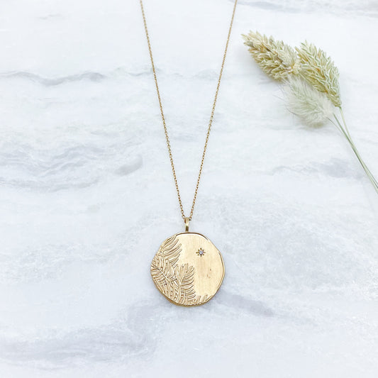 Long collier en chaîne maille forçat de 50 cm. Médaille de 2.6 cm de diamètre au contours irréguliers représentant de beaux feuillages et petit éclat scintillant en forme d'étoile. Or mat en partie recyclé couleur champagne