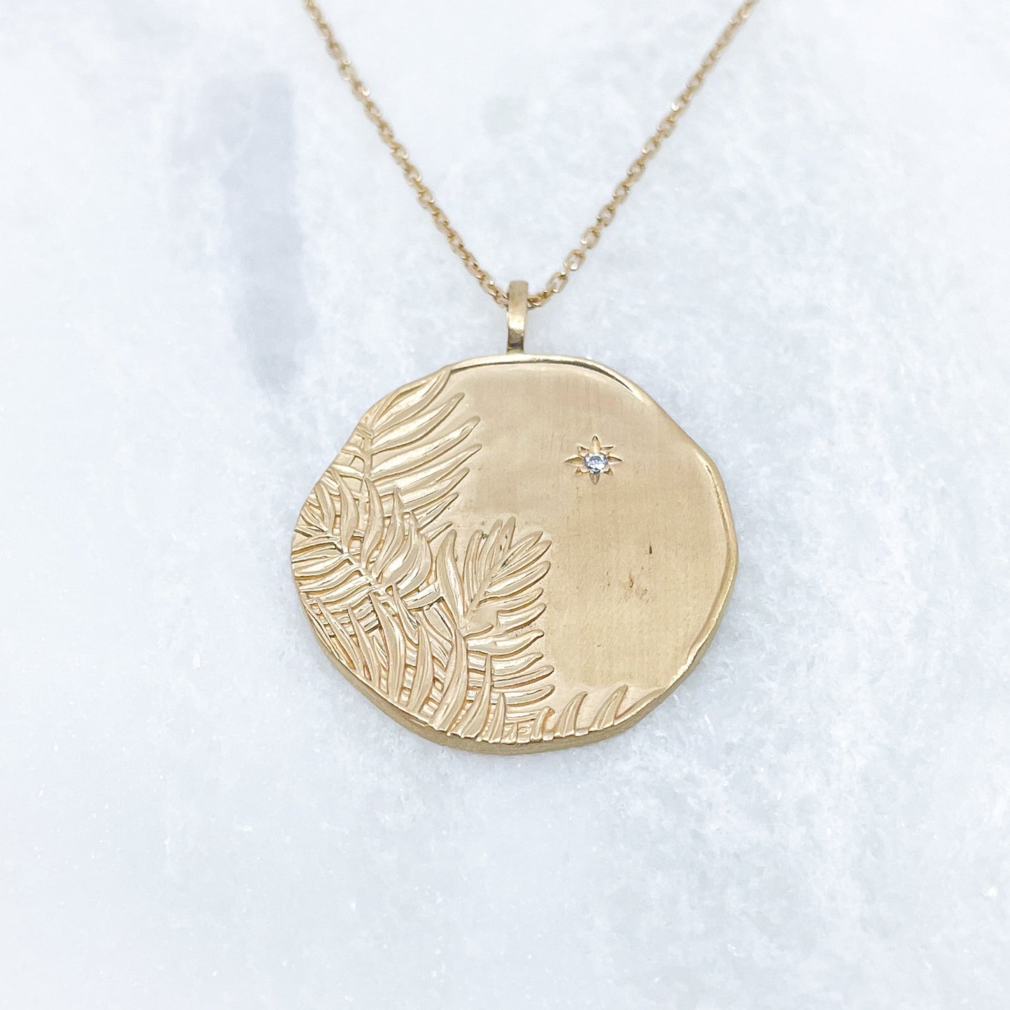 Long collier en chaîne maille forçat de 50 cm. Médaille de 2.6 cm de diamètre au contours irréguliers représentant de beaux feuillages et petit éclat scintillant en forme d'étoile. Or mat en partie recyclé couleur champagne