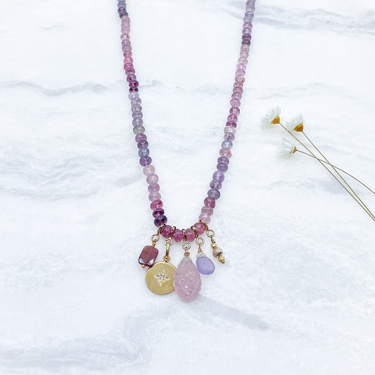 Collier long de 58 cm en pierres fines naturelles de spinelle dans des dégradés de rose à bleu gris. 5 charms : 3 pierres fines naturelles de teintes harmonieuses, une médaille ronde et un pendentif en petites perles plaqué or. Chaîne de réglage de 4 cm et fermoir plaqué or 18 carats