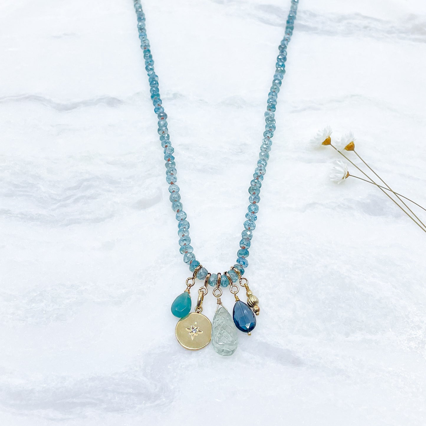 Collier long de 60 cm en pierres fines de zircon naturel d'un bleu azur. 5 charms : 3 pierres fines naturelles de teintes bleu-vert, une médaille ronde et un pendentif en petites perles plaqué or. Chaîne de réglage de 4 cm et fermoir plaqué or 18 carats