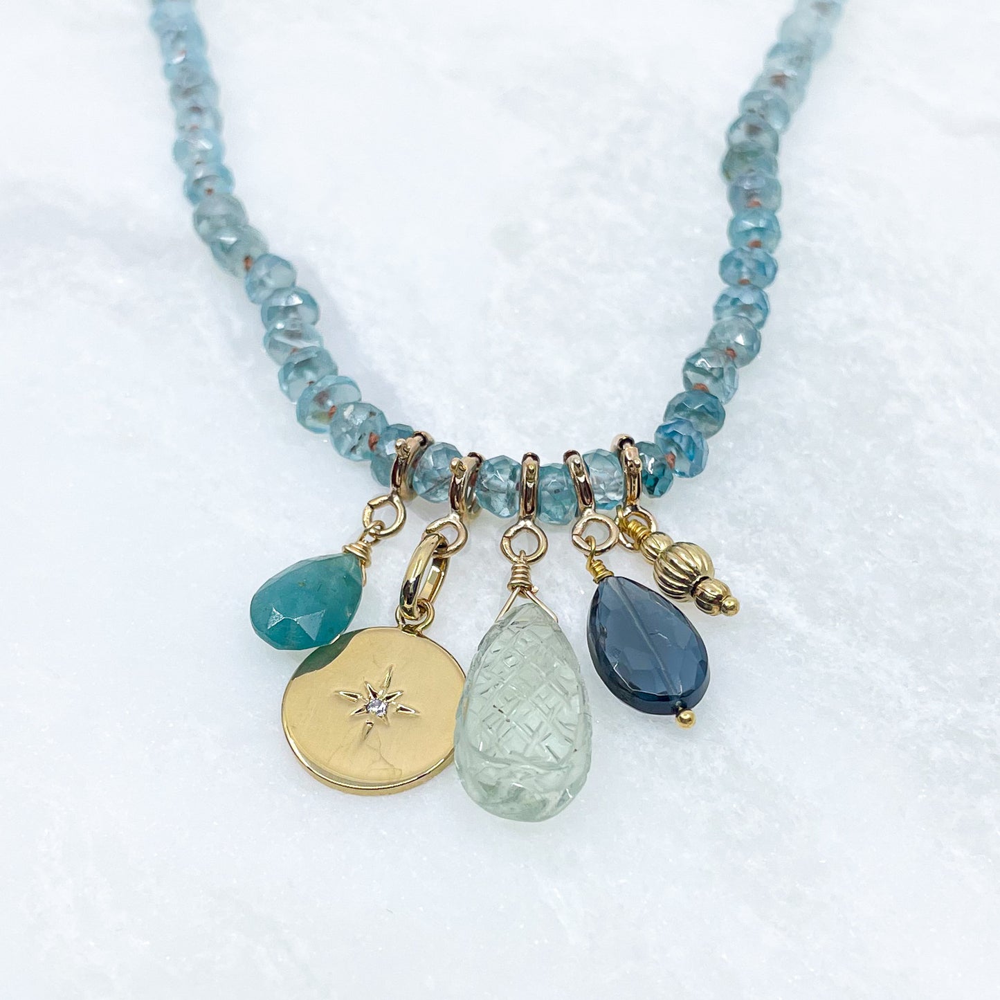 Collier long de 60 cm en pierres fines de zircon naturel d'un bleu azur. 5 charms : 3 pierres fines naturelles de teintes bleu-vert, une médaille ronde et un pendentif en petites perles plaqué or. Chaîne de réglage de 4 cm et fermoir plaqué or 18 carats